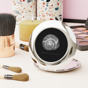 front view of a compact mirror that is black with a shell and writing underneath that says 'imperfect is perfect' on a desk with makeup 