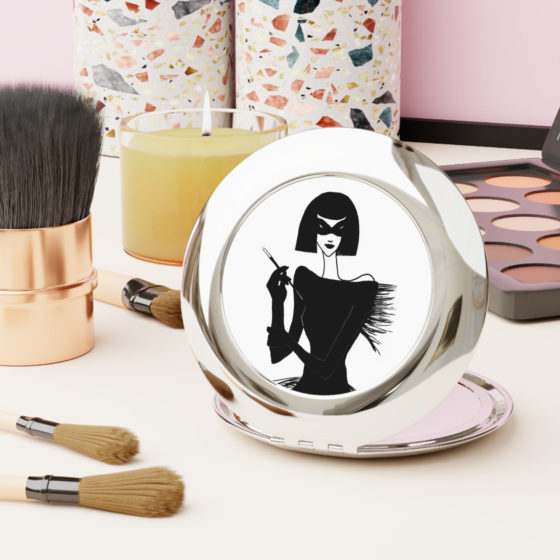 front view of a compact mirror on a desk with makeup with an ink drawing of a 1920's woman in a mask and holding a long cigarette