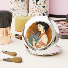 front view of a compact mirror with a painted image of a Geisha holding some violets on a desk with makeup